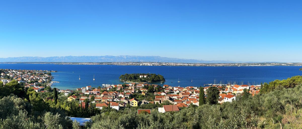 A view from a hill of a seaside village, its harbour and a small islet.